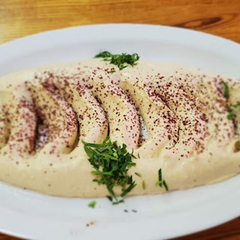 Creamy hummus with chicken slices, sprinkled with sumac and fresh parsley, on a white plate.